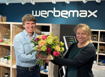 Gerhard Bezold Geschäftsführer werbemax erteilt Prokura Irina Belger im werbemax Showroom mit Blumenstrauß