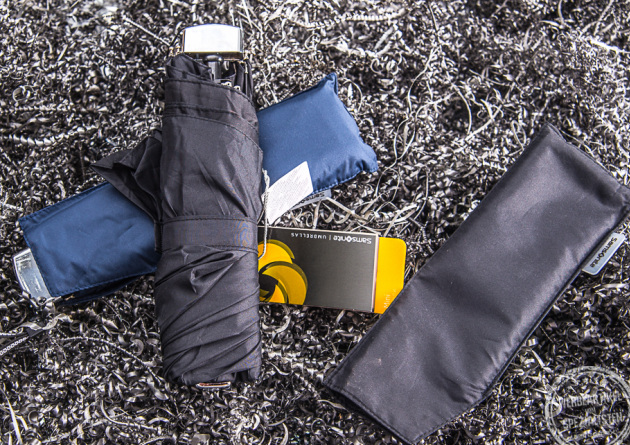 Schwarzer Mini Taschenschirm liegt auf dem blauen Pendant dazu. Die H&uuml;lle des schwarzen Schirms liegt daneben.