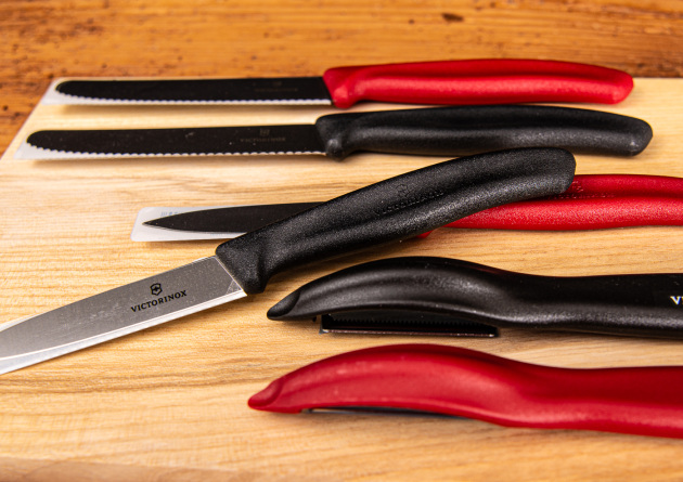 Nahaufnahme von Victorinox Gemüsemesserset plus Sparschäler mit roten und schwarzen Griffen, liegend auf Holzbrett.