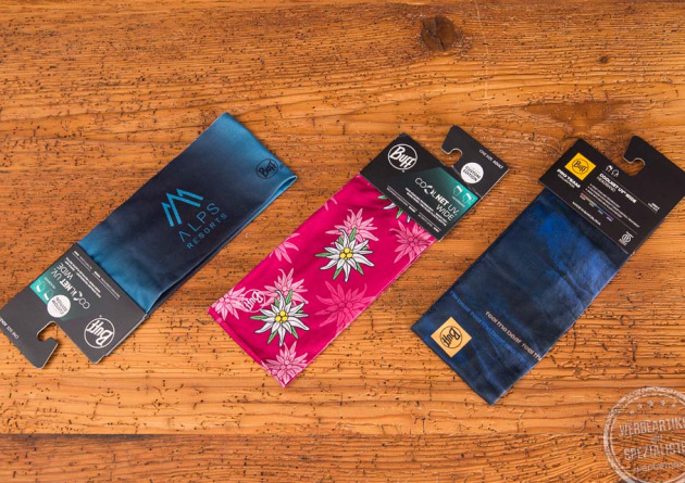 UV Coolnet Headband von BUFF mit individuellem Design. Auf dem Holztisch sieht man 3 Beispiele. 2 in Blau mit dezentem Logodruck und 1x in Pink mit Edelweiß