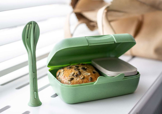 Grüne Lunchbox geöffnet und gefüllt mit einem Brötchen und einem kleinen Gefäß für Soßen plus Besteck als Werbeartikel.