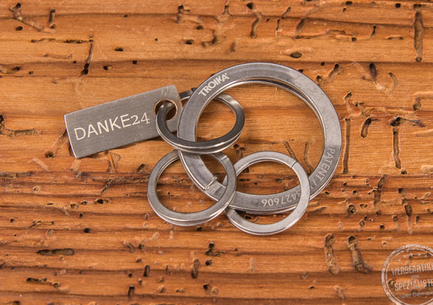 Ein Troika Schlüsselanhänger Nail Guard liegend auf Holzfläche mit Gravurplättchen und Aufdruck Danke.