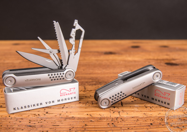 Zwei Taschenmesser Richartz Struktura Knife 15+ auf Holzoberfl&auml;che. Ein Tachenmesser ist ge&ouml;ffnet und liegt auf der Verpackung, das andere Tachenmesser ist geschlossen liegend auf der Verpackung. 