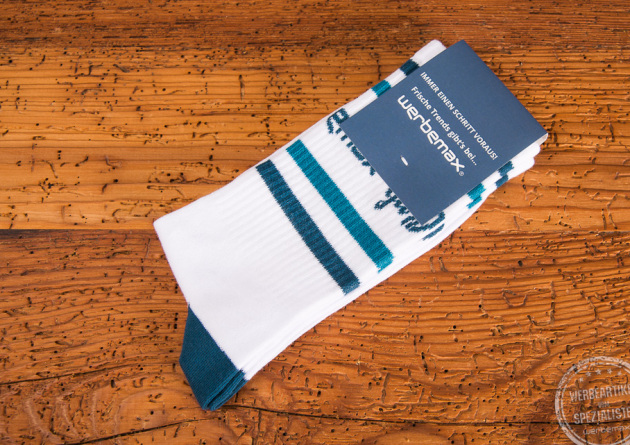 Werbesocken für die Firma werbemax - weiße Tennissocken mit petrolfarbenen Einwebungen an der Ferse sowie dem Bund. Besonders hochwertig wirkt das Giveaway dank der papierbanderole oben, die ebenfalls in petrol gestaltet ist