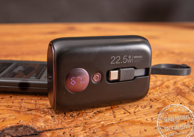 Eine schwarze Powerbank mit Ladestandsanzeige als Werbeartikel und einem Handy im Hintergrund.