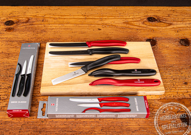 Zwei Victorinox Gemüsemesser Sets inkl Schäler mit den Grifffarben rot und schwarz in Verpackung und auf einem Brett liegend.