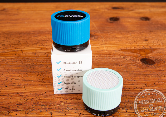 Ein schwarzer und ein wei&szlig;er Bluetooth Speaker mit buntem Gummiring als Merch auf Holzplatte.