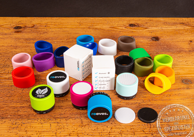 Mehrere Bluetooth Lautsprecher mit verschiedenen bunten Gummringen auf einer Holzplatte als Werbegeschenk.