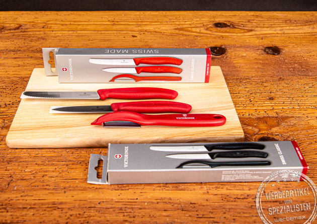 Zwei Gemüsemesser-Set Verpackungen von Victorinox liegend auf einem und neben einem Holzbrett. Ein Gemüsemesser Set mit Sparschäler mit rotem Griff liegend auf dem Holzbrett.