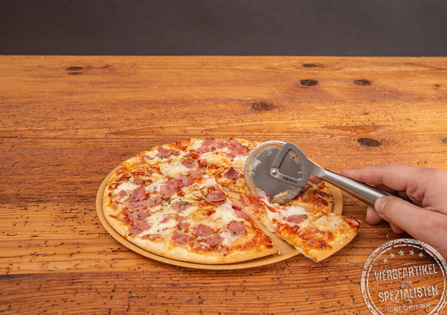 Hochwertiger Pizzaroller mit Lasergravur als Mitarbeitergeschenk auf Holztisch. Pizzaroller schneidet eine frische Pizza. 