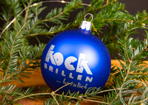 Blaue Weihnachtskugel mit weißem Logo, dekoriert an Tannenzweigen – personalisierte Christbaumkugel als festliches Kundengeschenk zu Weihnachten und elegante Eventdekoration.