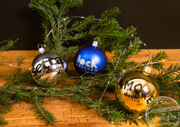 Personalisierte Weihnachtskugeln in Silber, Blau und Gold mit Logo, dekoriert an Tannenzweigen auf Holzplatte – individuelle Christbaumkugeln als Kundengeschenk zu Weihnachten.