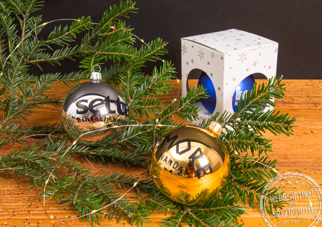 Silberne und goldene Weihnachtskugeln mit Logo, dekoriert mit Tannenzweigen und Geschenkbox – personalisierte Glaskugeln als festliche Werbegeschenke und hochwertige Christbaumdekoration für Weihnachten.