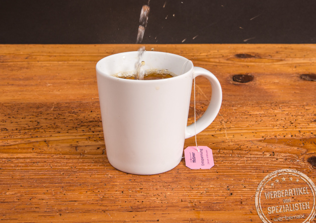 Wei&szlig;er SND Porzellanbecher mit Teebeutel, hei&szlig;em Wasser aufgegossen &ndash; ideal als Werbeartikel mit Logo.