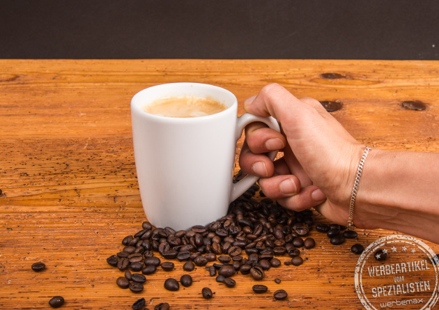 Person h&auml;lt SND Porzellanbecher mit Kaffee, umgeben von Kaffeebohnen &ndash; individueller Becher mit Logo m&ouml;glich.