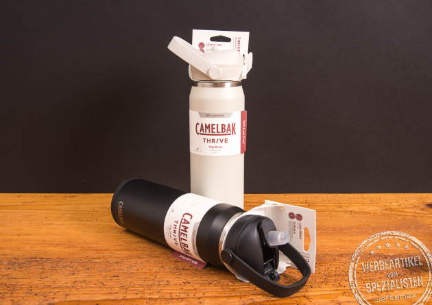 CamelBak Thrive Trinkflaschen in Weiß und Schwarz mit FlipStraw- und Chug-Verschluss – doppelwandig isolierte Edelstahlflaschen als nachhaltige Werbegeschenke mit Logo von werbemax auf Holztisch.