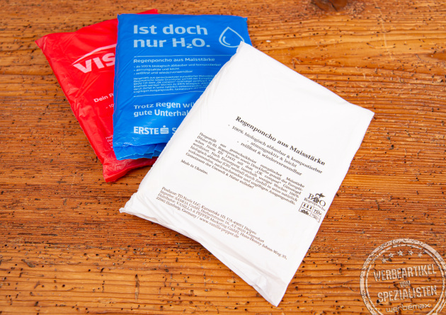 Mehrere farbige Verpackungen biologisch abbaubarer Regenponchos aus Maisstärke mit individuellen Logos – nachhaltige Regencapes als Werbeartikel und Festival-Giveaways von werbemax.