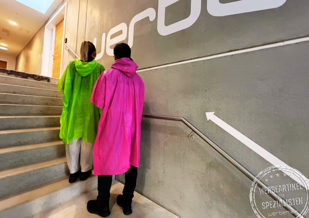 Zwei Personen tragen pinke und grüne Regenponchos aus biologisch abbaubarer Maisstärke und gehen eine Treppe hinauf – nachhaltige Regenponchos mit Logo als Werbeartikel für Festivals und Events von werbemax.