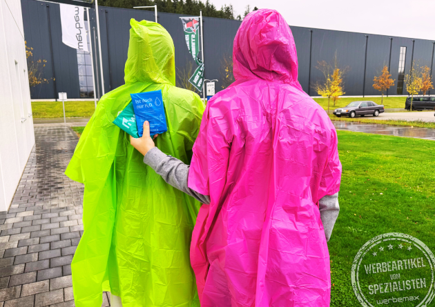 Zwei Personen mit grünen und pinken Regencapes aus biologisch abbaubarer Maisstärke gehen bei Regen – nachhaltige Regenponchos mit Logo als Werbeartikel für Festivals, Events und Tourismus von werbemax.