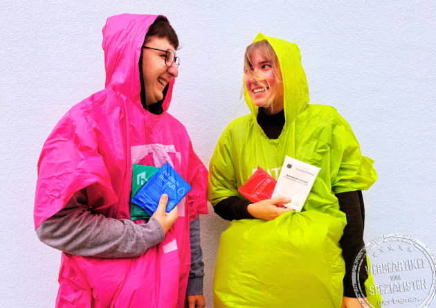 Zwei Personen tragen farbige Regenponchos aus biologisch abbaubarer Maisstärke mit Logo – nachhaltige Werbeartikel für Events, Festivals und Tourismusaktionen von werbemax.