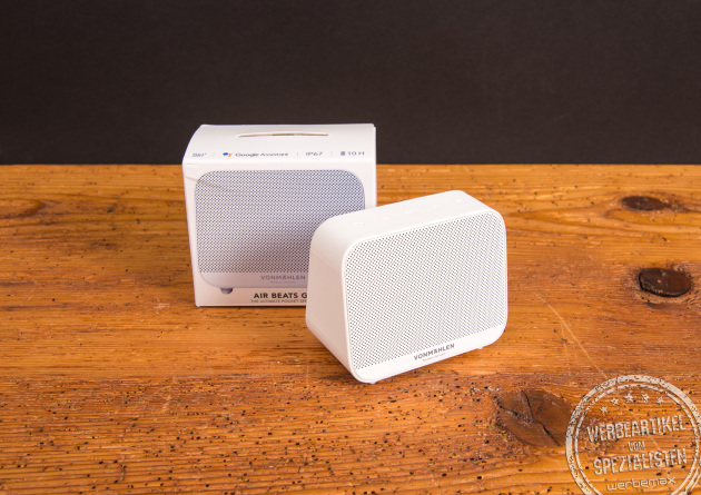 Bluetooth Box Air Beats Go in Weiß auf einem Holztisch stehend mit der Fachhandelsverpackung im Hintergrund. Der weiße Karton mit der Produktabbildung drückt nochmal Wertigkeit bei den Beschenkten aus. 