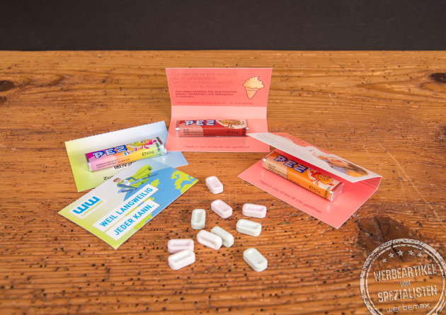 Verschiedene bedruckte PEZ-Werbekarten mit PEZ-Sticks als kreatives Give-away, pr&auml;sentiert mit Bonbons auf Holztisch.