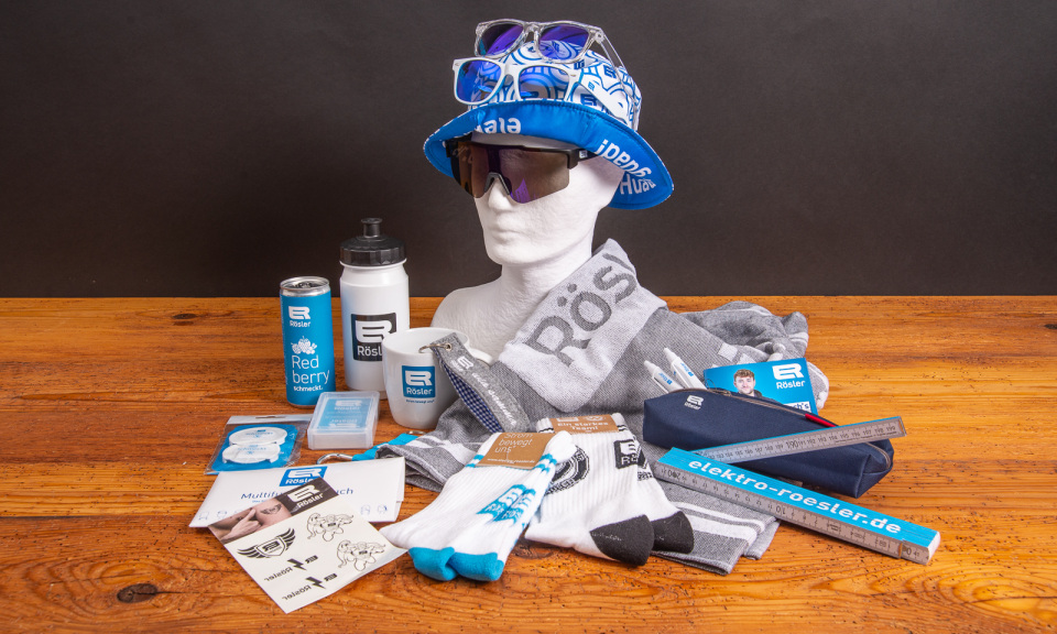 Merchandising-Set mit Rösler Werbeartikeln: bedrucktes Handtuch, Trinkflasche, Tasse, Socken, Sonnenbrillen, Bucket Hat, Lineal, Schreibwaren, Energy-Drink-Dose und weitere Sportfanartikel mit Logo auf Holzfläche arrangiert.