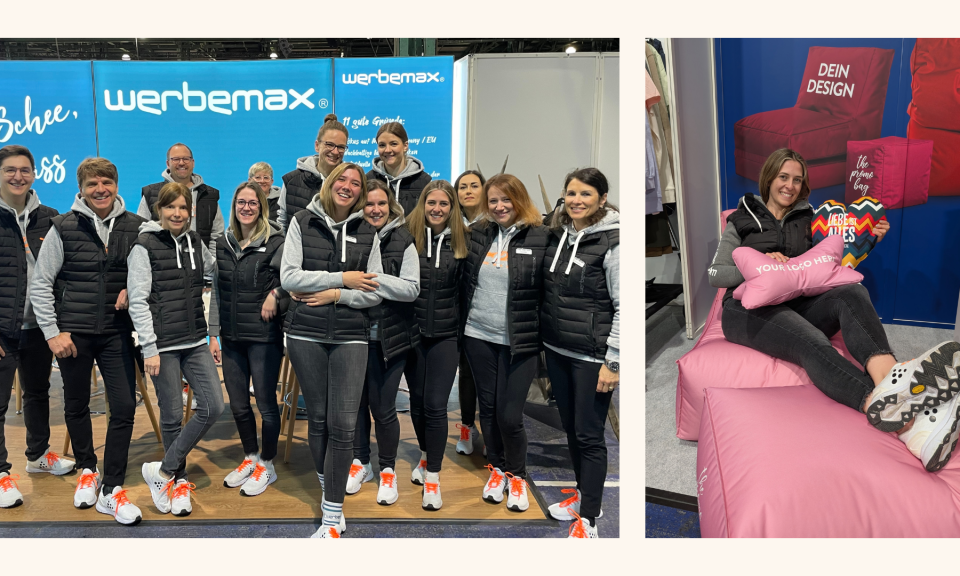 Teamfoto von werbemax auf der Werbeartikelmesse WerbeWelt: eine gro&szlig;e, fr&ouml;hliche Gruppe im einheitlichen Outfit mit schwarzen Westen, grauen Hoodies und wei&szlig;en Sneakern mit orangefarbenen Schn&uuml;rsenkeln vor blauem werbemax-Messestand; daneben eine Mitarbeiterin entspannt auf rosa Sitzs&auml;cken sitzend, umgeben von individuell bedruckbaren Kissen und Herz-Giveaways &ndash; Beispiel f&uuml;r personalisierbare Werbeartikel und modernes Messebranding.