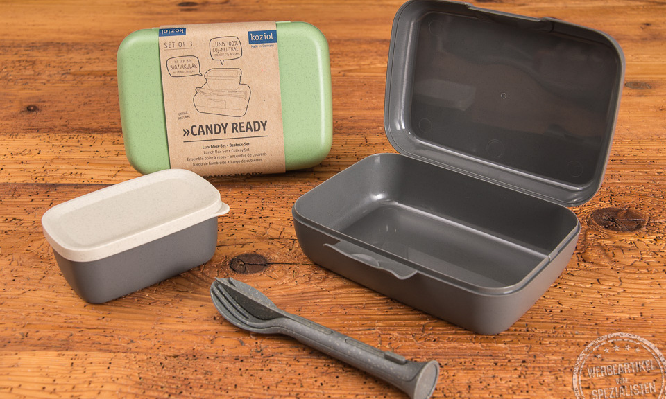 Ein geöffnetes graues koziol Candy Ready Lunchbox Set inklusive Besteck und kleiner auslaufsicheren Dose, sowie eine geschlossene grüne Candy-Ready Lunchbox. 