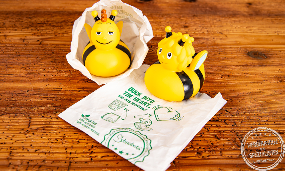 Quietscheenten Biene Maja und Biene Willi in Verpackungst&uuml;te auf Holztisch.