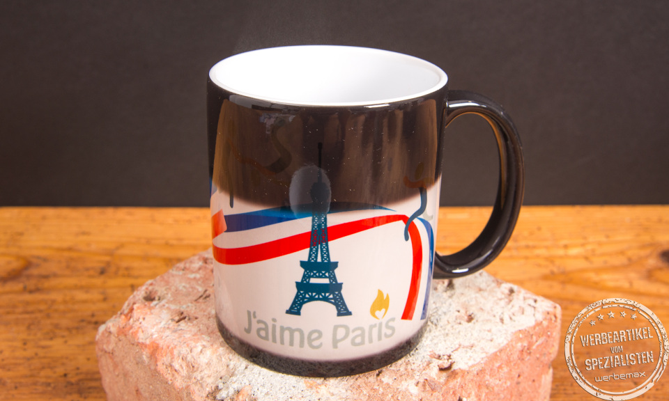 Thermotasse mit Farbwechsellack zeigt Eiffelturm-Motiv durch heiße Befüllung – Magic-Werbetasse von werbemax auf Backstein.