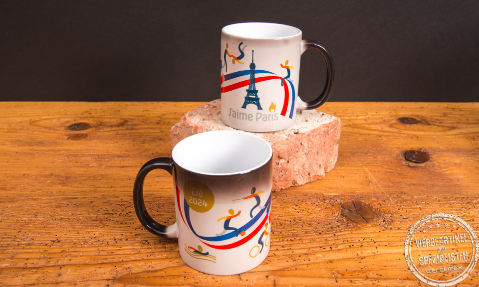 Zwei Magic-Tassen mit Farbwechsel-Design „J’aime Paris“ zeigen das Motiv durch heiße Flüssigkeit – bedruckte Werbeartikel Tassen mit Thermoeffekt von werbemax auf Backstein.