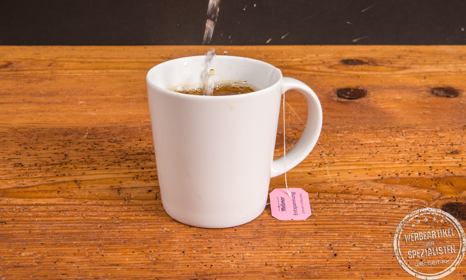 Weißer SND Porzellanbecher mit Teebeutel, heißem Wasser aufgegossen – ideal als Werbeartikel mit Logo.