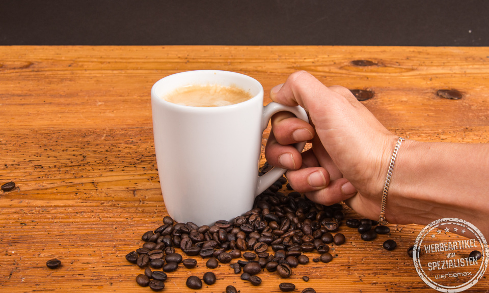 Person hält SND Porzellanbecher mit Kaffee, umgeben von Kaffeebohnen – individueller Becher mit Logo möglich.
