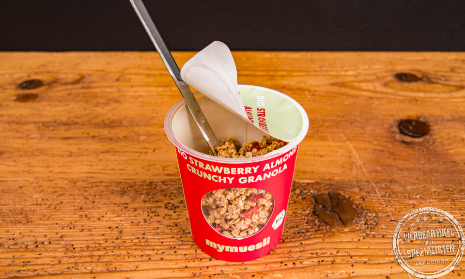 Einzelner mymuesli M&uuml;sli to go Becher auf Holztisch &ndash; idealer Werbeartikel f&uuml;r Unternehmen.