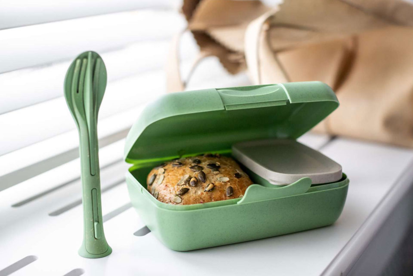 Grüne Lunchbox geöffnet und gefüllt mit einem Brötchen und einem kleinen Gefäß für Soßen plus Besteck als Werbeartikel.