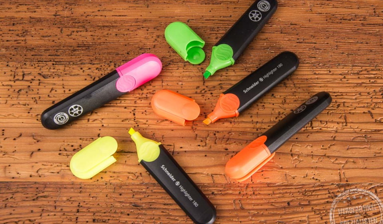 Vier Textmarker der Marke Schneider in Schwarz mit pinkfarbener, gelber, orangefarbener und grüner Kappe. Geöffnet auf einer Holzunterlage