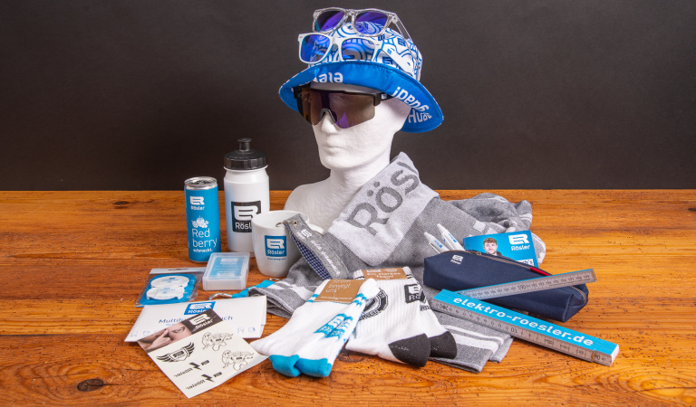 Merchandising-Set mit R&ouml;sler Werbeartikeln: bedrucktes Handtuch, Trinkflasche, Tasse, Socken, Sonnenbrillen, Bucket Hat, Lineal, Schreibwaren, Energy-Drink-Dose und weitere Sportfanartikel mit Logo auf Holzfl&auml;che arrangiert.