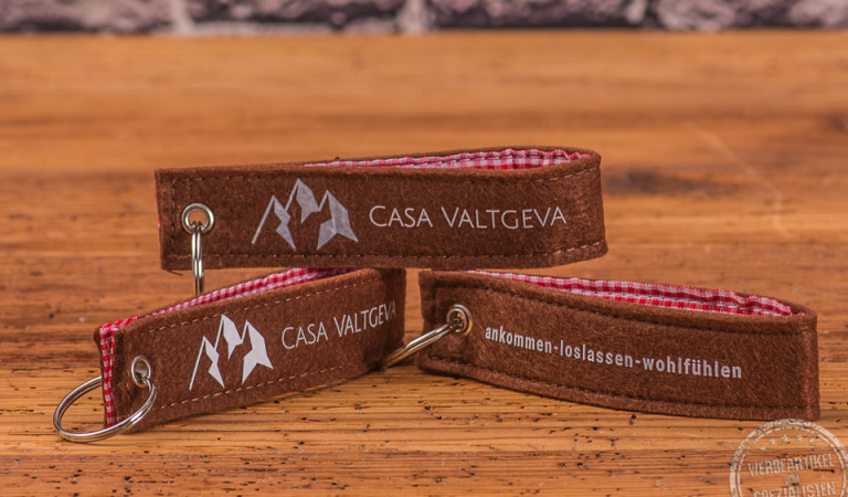 Vorne und hinten bestickter, brauner Filzschlüsselanhänger mit rotkariertem Stoff innen als Werbegeschenk für Kunden und Mitarbeiter auf Holzplatte