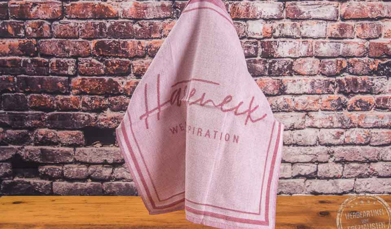 Umsetzungsbeispiel Logo auf rotem Geschirrtuch als Werbegeschenk für Kunden hängend vor Steinmauer