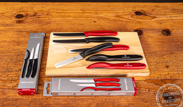 Zwei Victorinox Gemüsemesser Sets inkl Schäler mit den Grifffarben rot und schwarz in Verpackung und auf einem Brett liegend.