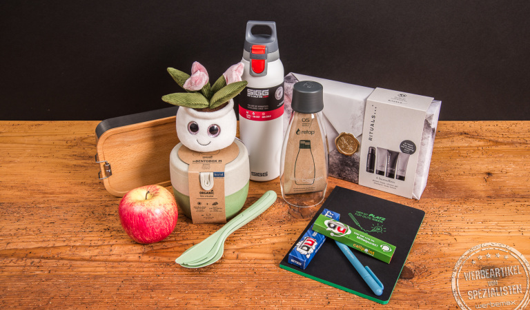 Moderne nachhaltige Werbegeschenke für Mitarbeiter – liebevoll zusammengestellte Geschenkideen wie SIGG Trinkflasche, Retap Bottle, Notizbuch, Bento-Box und Rituals Pflege-Set auf Natur-Holztisch.