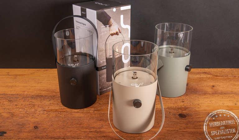 Cosiscoop Original Gaslaternen in Schwarz, Beige und Olivgrün mit Verpackung auf Holztisch – hochwertiges Outdoor-Licht als Werbegeschenk mit Logo