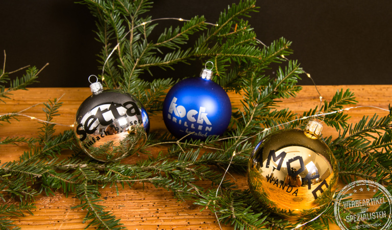Personalisierte Weihnachtskugeln in Silber, Blau und Gold mit Logo, dekoriert an Tannenzweigen auf Holzplatte &ndash; individuelle Christbaumkugeln als Kundengeschenk zu Weihnachten.