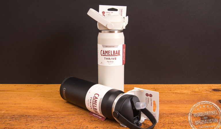 CamelBak Thrive Trinkflaschen in Weiß und Schwarz mit FlipStraw- und Chug-Verschluss – doppelwandig isolierte Edelstahlflaschen als nachhaltige Werbegeschenke mit Logo von werbemax auf Holztisch.