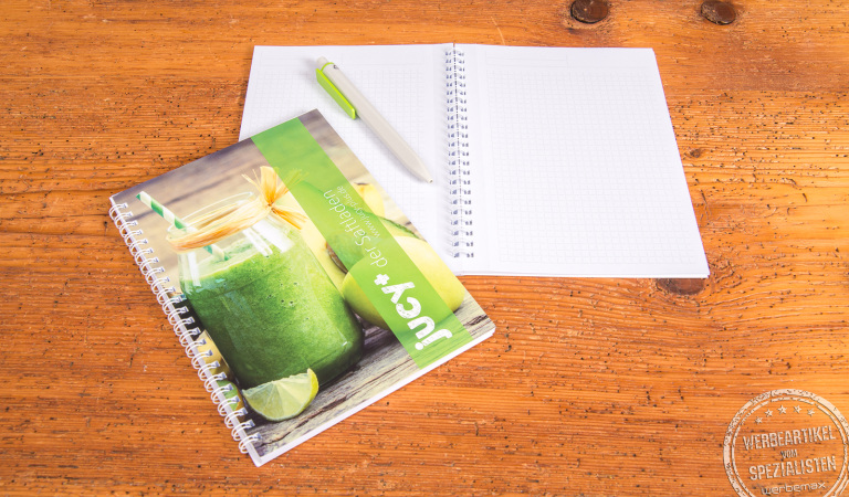 G&uuml;nstiges A5 Notizbuch mit Spiralbindung und individualisiertem Titelblatt, ideal als Werbeartikel oder Kundengeschenk. Abgebildet auf Holztisch mit passendem Stift und aufgeschlagenen Seiten.