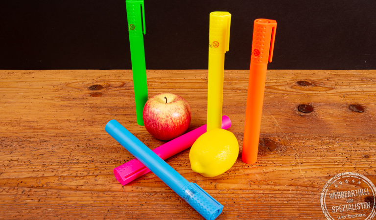 Jumbo Textmarker im XL-Format als Riechstifte in leuchtenden Farben auf Holztisch, dekoriert mit Apfel und Zitrone – kreative Werbeartikel und auffällige Giveaway-Idee für Messen und Promotion-Aktionen.