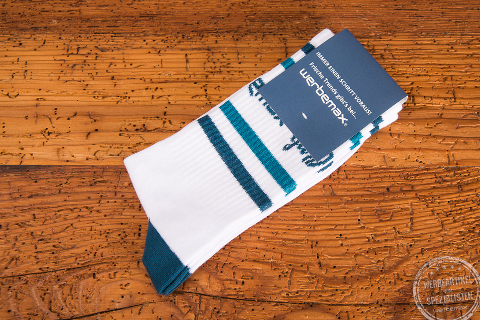 Werbesocken f&uuml;r die Firma werbemax - wei&szlig;e Tennissocken mit petrolfarbenen Einwebungen an der Ferse sowie dem Bund. Besonders hochwertig wirkt das Giveaway dank der papierbanderole oben, die ebenfalls in petrol gestaltet ist